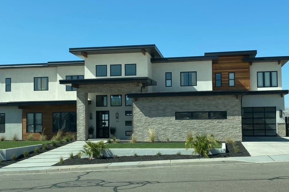 Two-story modern home with smooth off-white stucco, gray ledgestone on the entry column and lower front wall, and wood-tone accent panels